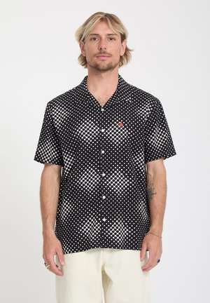 Homme blond portant une chemise noire à manches courtes à boutons, ornée de motifs carrés blancs et de points, associée à un pantalon de couleur claire.