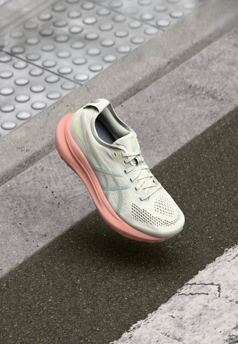 Chaussure de course légère en crème pâle avec semelle rose, présentant un dessus en mesh, une boîte à orteils perforée et de subtils accents bleu sur les côtés.