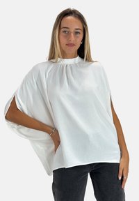 Haut blanc à manches courtes et col montant, avec une texture douce et un éclat subtil ; doté d'un design fluide et ample avec des fentes latérales. Porté avec un jean foncé.