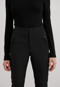 Svarte tettsittende bukser med glatt tekstur, med to glidelåslommer på sidene, dobbel knappelukking og kontrasterende kant langs sidene.