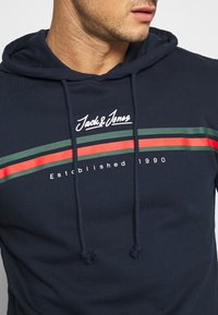 Felpa blu navy con cordini, caratterizzata da strisce orizzontali verdi e rosse e logo "Jack & Jones Established 1990" sul petto.