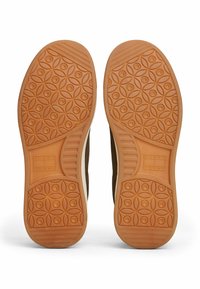 Brązowa gumowa podeszwa z teksturowanym wzorem z okrągłymi wzorami. W centrum widoczna etykieta z logo. Gładkie beżowe detale po bokach.