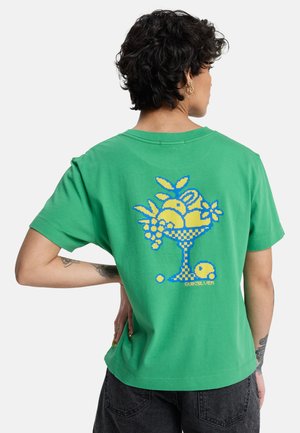 Personne aux cheveux courts et bouclés portant une chemise verte avec un motif pixel art représentant un saladier de fruits jaune et bleu dans le dos.