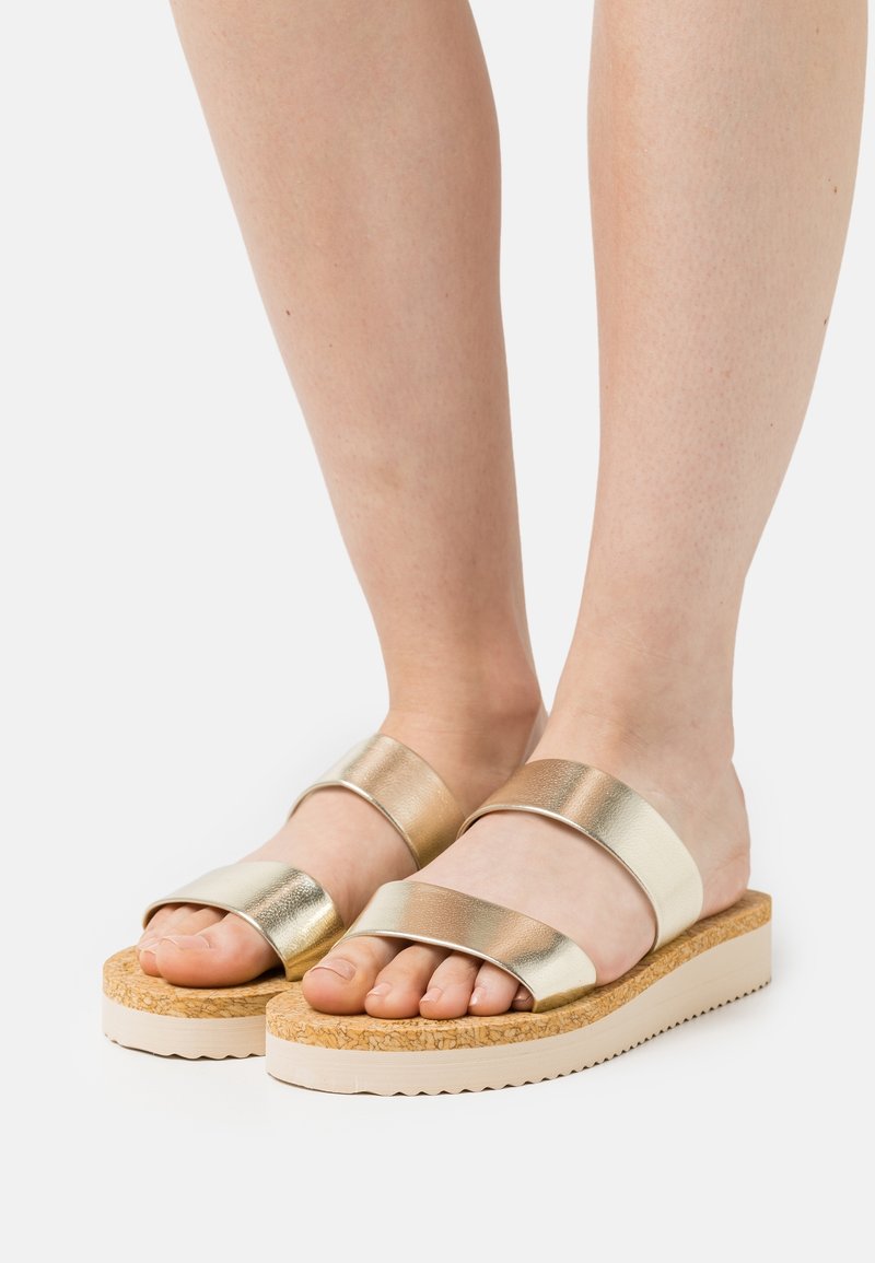 Pieds portant des sandales métalliques dorées à enfiler avec deux larges brides et une semelle plateforme texturée liège sur fond blanc uni.