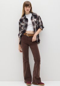 Bruine corduroy flare-pants gecombineerd met een witte crop top en een geruite flanellen overhemd. Schoenen zijn kastanjebruin met een beige zool.