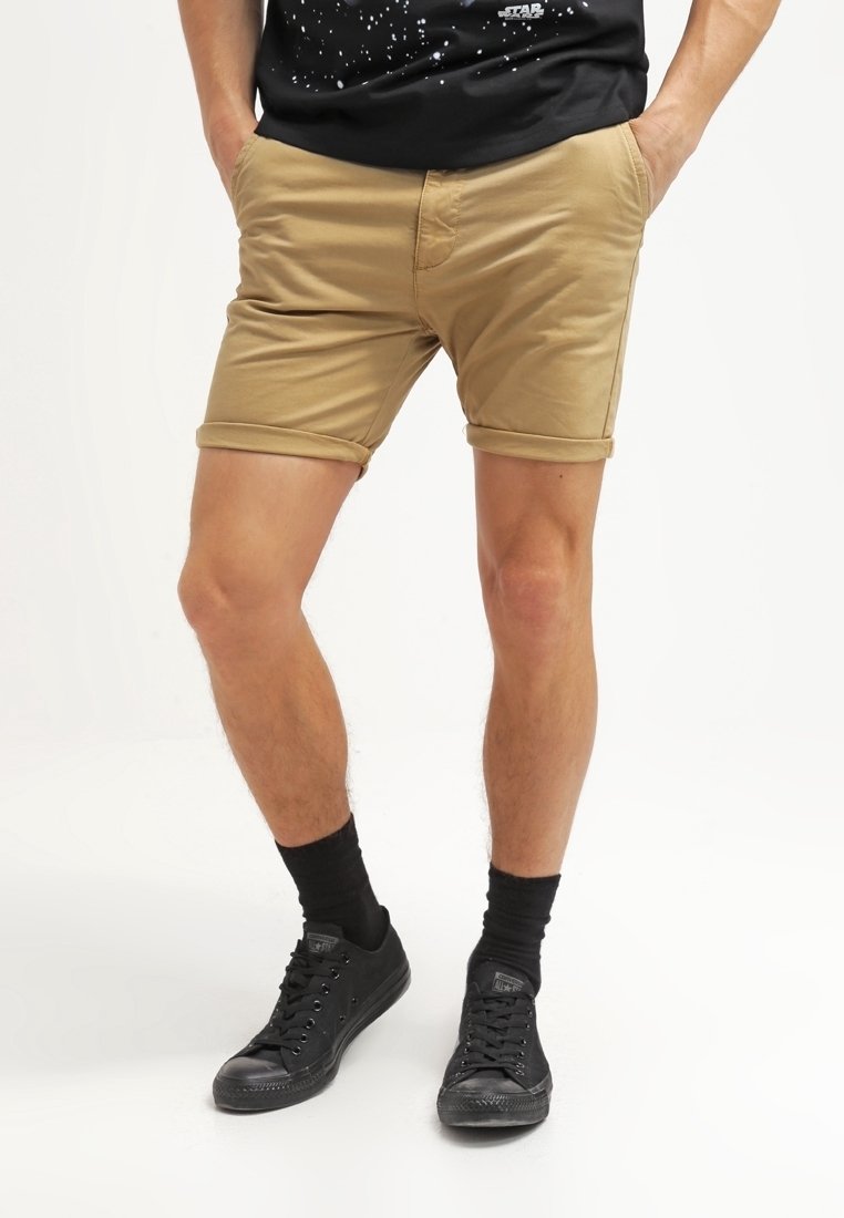 Homme portant un short kaki retroussé, des chaussettes noires montantes et des baskets noires, debout les mains dans les poches sur un fond blanc.