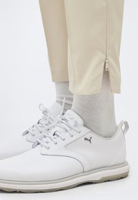 Λευκά παπούτσια Puma με κορδόνια, φορεμένα με ανοιχτό γκρι κάλτσες και μπεζ παντελόνια με μικρό φερμουάρ πάνω από τον αστράγαλο.