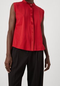 Blusa rossa senza maniche con colletto alla mandarino, chiusura frontale con bottoni e leggera forma ad A. Tessuto liscio, indossata sopra pantaloni neri.