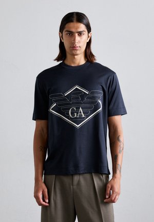 T-shirt en coton bleu marine à manches courtes avec un grand graphique géométrique blanc comportant les lettres "GA". Design épuré, coupe décontractée.