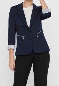 Marinblå blazer med skräddarsydd passform, två framfickor med dragkedja och vikta randiga manschetter. Slät tygkvalitet, en knappstängning och strukturerade slag.