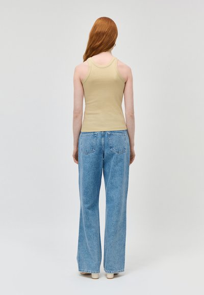 Personne aux longs cheveux roux portant un débardeur beige et un jean bleu clair taille haute à jambes larges, debout de dos sur un fond uni.