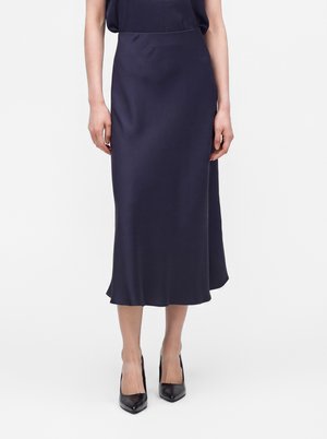 Mujer con una falda midi azul marino y tacones altos negros de punta afilada, de pie frente a un fondo claro y liso.