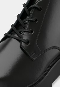 Čierna lesklá kožená topánka s okrúhlou špičkou, zložitým prešívaním a šiestimi dierkami na šnúrky. Hladký povrch s elegantným lesklým finišom.