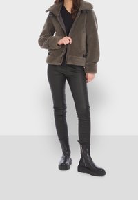 Femme portant une veste marron en matière pelucheuse avec fermeture éclair, un pantalon en cuir noir et des bottes en cuir noir avec des fermetures éclair latérales, debout sur un fond gris.