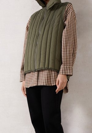 Personne portant une veste matelassée sans manches vert olive par-dessus une chemise à carreaux marron et blanche à manches longues et un pantalon noir, debout contre un mur clair.