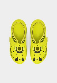 Φωτεινά κίτρινα παπούτσια τύπου clog με τρύπες αερισμού και εσωτερικές σόλες με μαύρες ρίγες, φωτογραφημένα από πάνω σε λευκό φόντο.