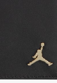 Czarna skórzana powierzchnia o teksturowanym wykończeniu, z wyraźnie widocznym złotym logo Jumpman. Gładkie i błyszczące akcenty.