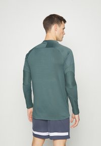 Maglia sportiva a maniche lunghe in verde scuro, caratterizzata da un design strutturato, tessuto liscio e dettagli testurizzati sulle maniche. Abbinata a pantaloni corti.
