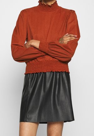 Langarmtop in roestkleur met een geplooide col en een elastische taille, gecombineerd met een zwarte leren minirok. Soepele textuur.