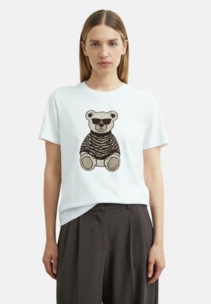 Frau trägt ein weißes T-Shirt mit einem Teddybär in Sonnenbrille und einem gestreiften Pullover aufgedruckt vorne, kombiniert mit dunklen, weiten Hosen.