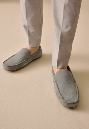 Person iført lysegrå bukser og grå ruskind slip-on loafers på et beige gulv.