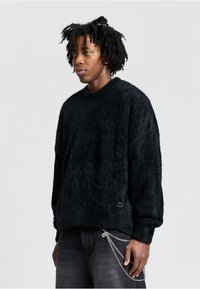 Schwarzer, flauschiger Pullover mit Rundhalsausschnitt, langen Ärmeln und seitlichem Reißverschluss. Kombiniert mit dunklen Jeans, die mit einer silbernen Kettenakzent versehen sind.