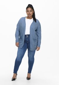 Világoskék kötött kardigán nyitott elülső résszel, bordázott mandzsettával és két elülső zsebbel, kék denim skinny farmernadrággal és fekete magassarkúval párosítva.