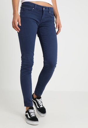 Osoba nosząca granatowe jeansy typu slim fit i czarno-białe sneakersy, stojąca na jednolitym jasnym tle.