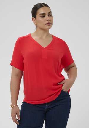 Kobieta o ciemnych włosach uczesanych w niski kok, ubrana w czerwoną bluzkę z krótkim rękawem i dekoltem w serek oraz ciemnoniebieskie jeansy, z jedną ręką w kieszeni.