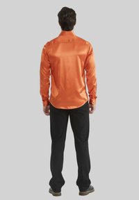 Chemise à manches longues en satin orange avec une finition lisse, poignets à bouton et ourlet arrondi, associée à un pantalon noir et des chaussures brunes.