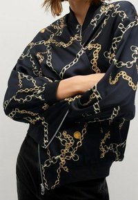 Chaqueta bomber negra con patrón de cadenas doradas, presentando un cierre con cremallera, puños acanalados y un detalle de logo redondo en el hombro.