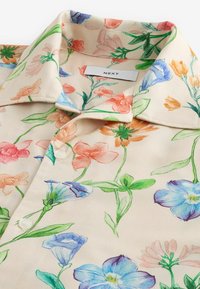 Chemise crème avec col orné d'un imprimé floral coloré dans des tons de bleu, orange, rose et vert, avec des boutons blancs et une étiquette "NEXT" à l'intérieur du col.