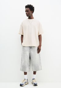 Beige, oversized pamut póló rövid ujjakkal, világosszürke farmer sorttal párosítva. A lábbelik között kék és bézs színű, vastag talpú sportcipők találhatók.