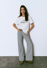 Camiseta blanca con gráfico negro de "Sports Club", combinada con pantalones de campana a rayas grises y blancas y zapatos de slip-on marrones con tachuelas.