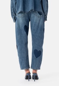 Jeans di denim blu con toppe a forma di cuore in una tonalità più scura, orlo sfrangiato, vestibilità rilassata, abbinati a scarpe con tacco grigie.