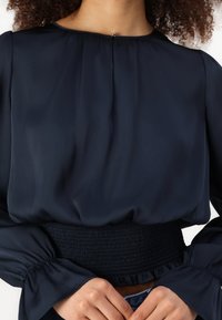 Temno modra bluza z nabranim okroglim ovratnikom, nabrano pasom in zvonastimi rokavi. Gladka tkanina z rahlim sijajem. Gumbasta zaponka na hrbtni strani.