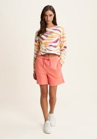 Top a maniche lunghe in un motivo ondulato multicolore abbinato a pantaloni corti color corallo con un elastico in vita; sneakers bianche completano l'outfit.