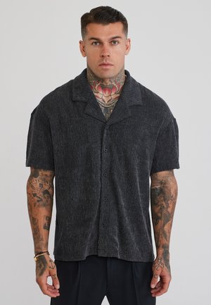 Mann mit Tattoos am Hals und an den Armen, der ein schwarzes kurzärmeliges Hemd und schwarze Hose vor einem einfarbigen grauen Hintergrund trägt.