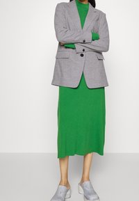 Blazer gris sur une robe verte côtelée, avec des manches longues et une jupe mi-longue. Chaussures gris clair à enfiler avec une surface texturée.