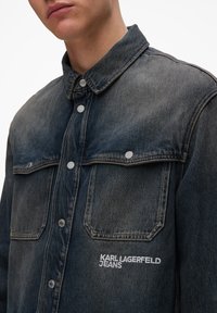 Mørkeblå denimjakke med knappestolpe foran, to brystlommer og brodert "KARL LAGERFELD JEANS"-logo på venstre lomme.