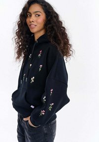 Černá mikina s kapucí s barevnou květinovou výšivkou, klokáním kapsou, nastavitelným stahovacím šňůrkou a volným střihem. Texturovaná tkanina s hladkým povrchem.