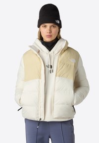 Puffjacka i kräm- och beige, med dragkedja fram, hög krage och sidofickor. Bärs över en ljus hoodie och matchas med marinblå byxor.