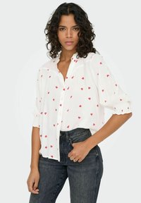 Camisa blanca de botones con patrones de corazones rojos, de tela ligera, con mangas fruncidas y un corte relajado, combinada con jeans de mezclilla oscura.