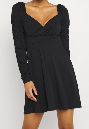 Kobieta w czarnej sukience z długimi rękawami z marszczeniami i dekoltem w kształcie litery V, stojąca na tle jasnego, jednolitego tła.