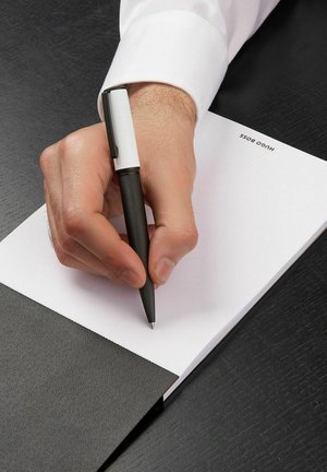 Un stylo noir texturé tenu dans une main, écrivant sur un carnet blanc. Le carnet a une couverture noire et présente "Hugo Boss" imprimé sur son bord.