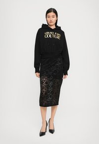 Svart hettegenser med gullbokstaver, kombinert med et svart blonde midiskjørt. Outfiten har en tettsittende silhuett og høyhælede sko.
