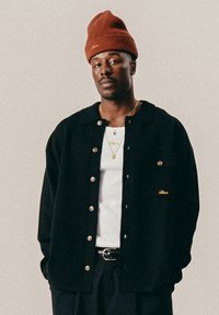 Man in een rode muts, met een zwarte overhemdjas over een witte shirt, versierd met gouden kettingen en hanger oorbellen, tegen een effen lichte achtergrond.