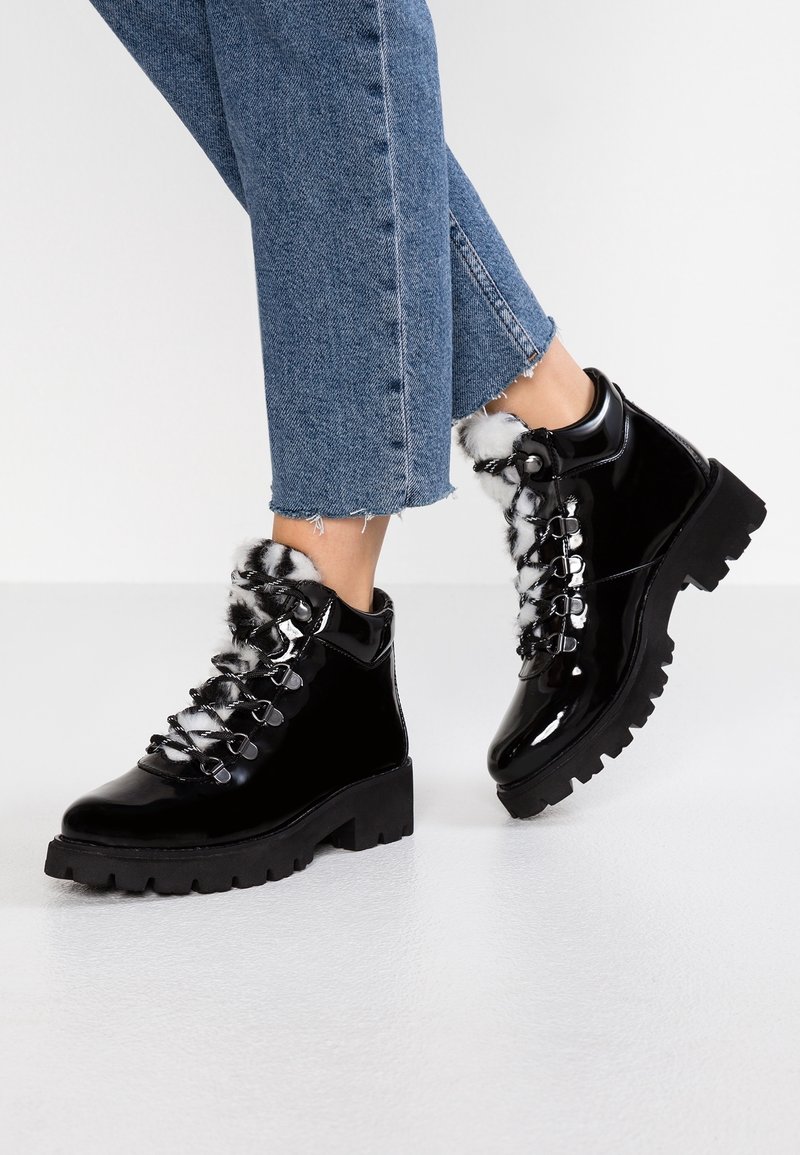Bottines en cuir verni noir avec bord en fausse fourrure, lacets à l'avant et semelles en caoutchouc épaisses, ornées d'œillets et d'accents métalliques.