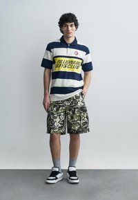 Fiatal férfi áll, egyik kezét a zsebében tartja, csíkos "Billionaire Boys Club" pólót visel, terepmintás cargo rövidnadrágot, szürke zoknit és fekete-fehér sportcipőt.