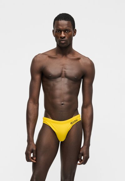 Άνδρας μοντέλο με σκούρο δέρμα που φοράει κίτρινα μαγιό χαμηλής μέσης με μαύρο κείμενο στον αριστερό γοφό, στέκεται μπροστά σε ένα απλό φωτεινό φόντο.